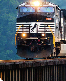 A black freight train engine with the number 9841 travels on a railway track, surrounded by a green, forested area. The front lights are on, and a horse logo is visible on the front